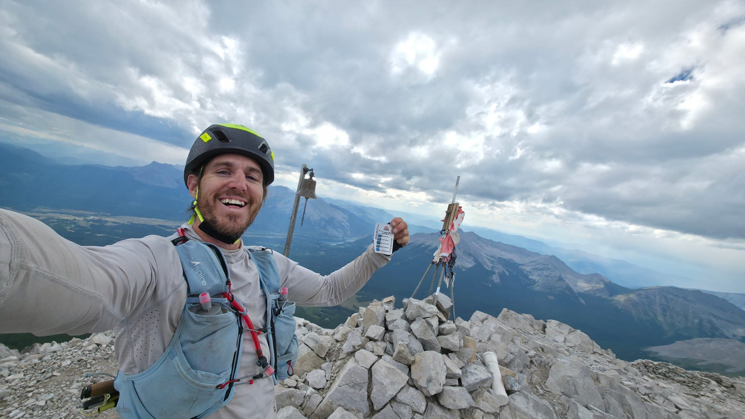 Shep on the summit of Crowsnest Mountain in a climbing helmet, holding up his punched High Five card beside the summit bell